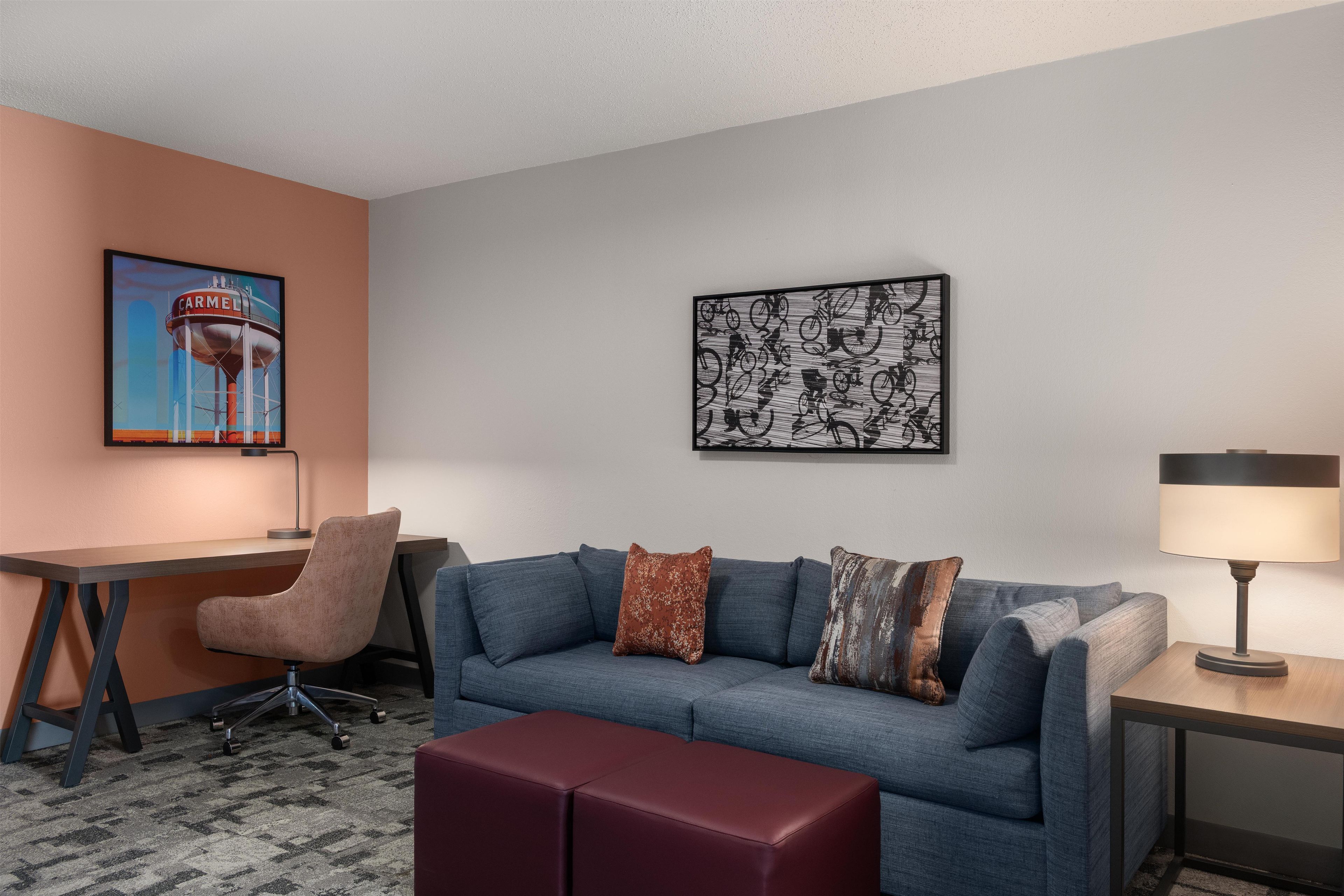 A hotel room with a desk, a blue sofa with cushions, a red ottoman, a lamp, and two wall art pieces.
