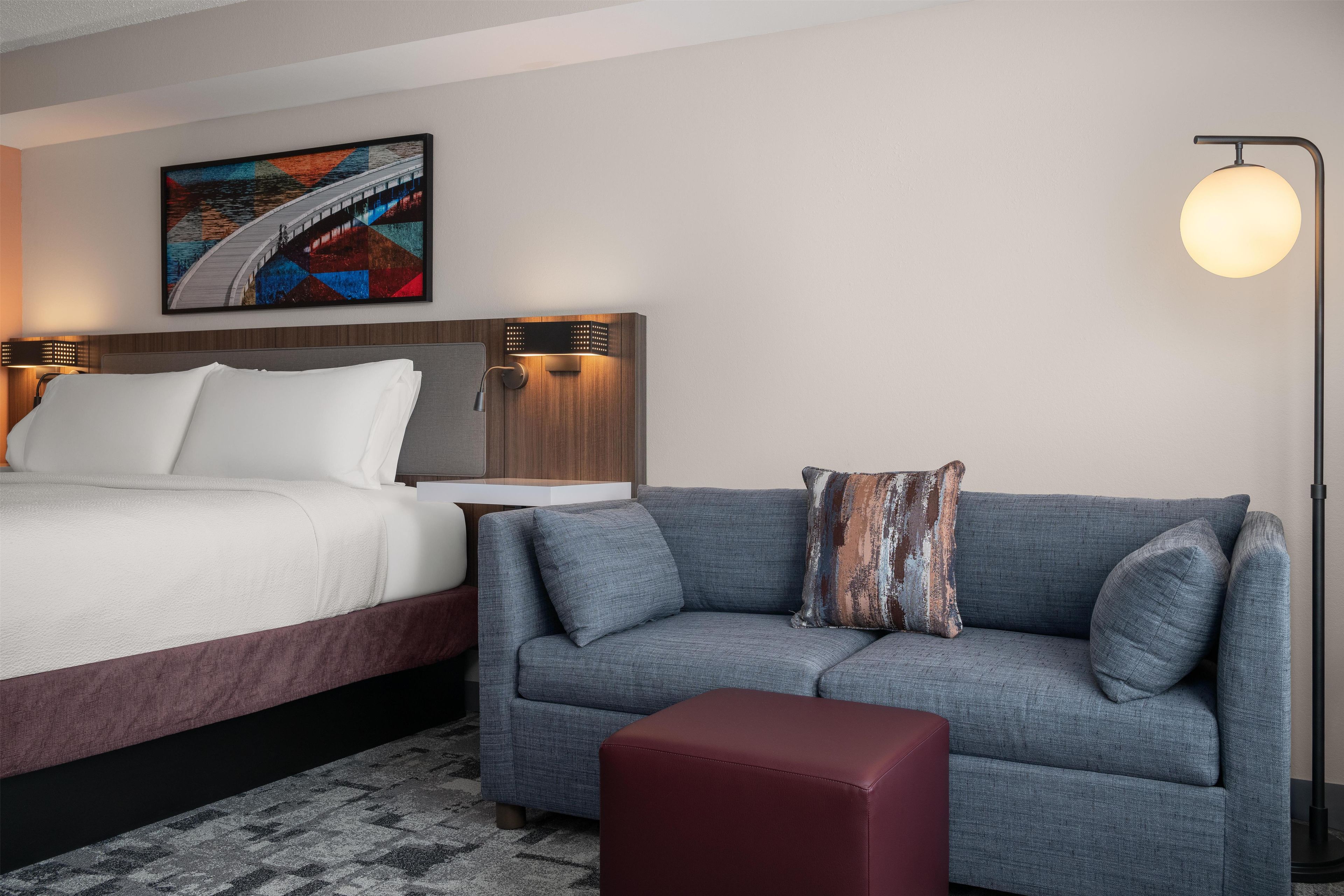 Ein Hotelzimmer mit Bett, einer Sitzecke, einem roten Hocker, einer Stehlampe und einem Gemälde an der Wand.