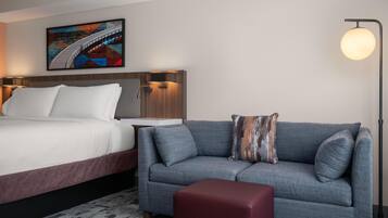 Ein Hotelzimmer mit Bett, einer Sitzecke, einem roten Hocker, einer Stehlampe und einem Gemälde an der Wand.