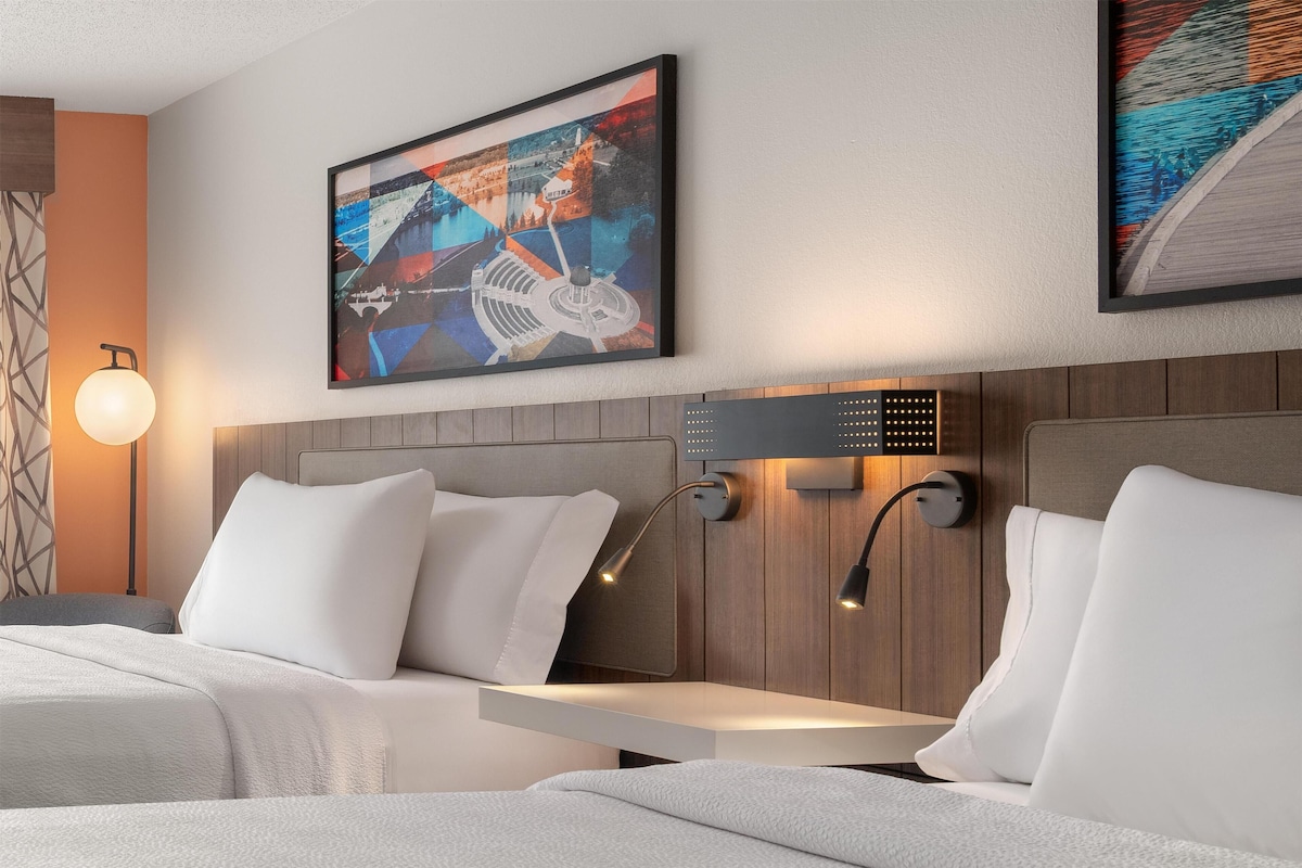 A modern hotel room with two beds, a wooden headboard, wall-mounted artwork, and a bedside table with a lamp.