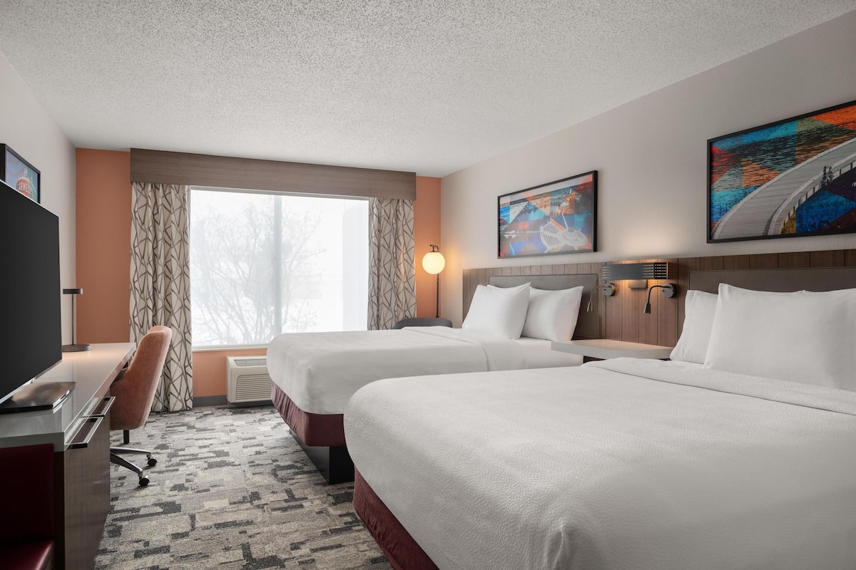 A hotel room with two beds, a desk, a chair, a TV, and a large window with curtains.