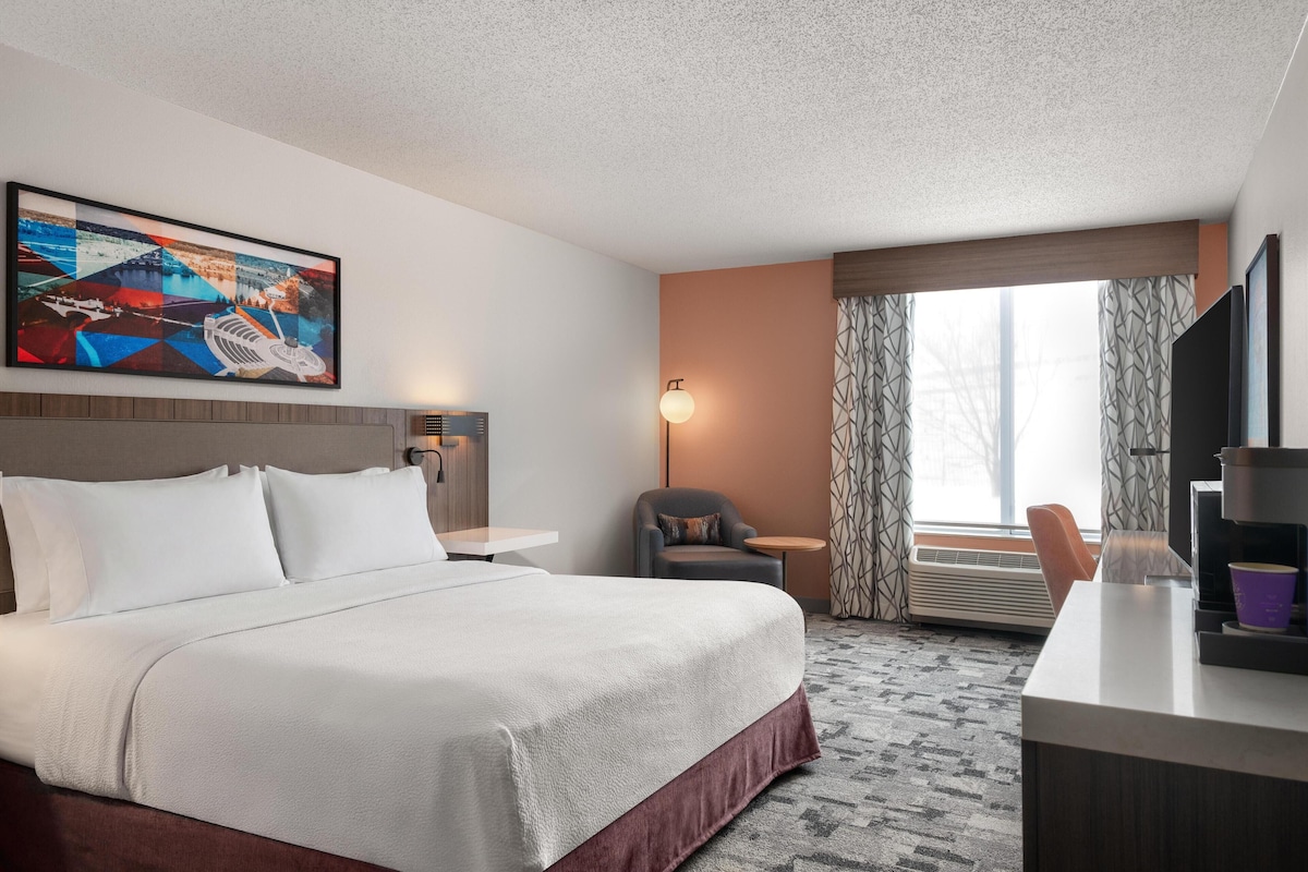 A hotel room with a large bed, a desk, a chair, a painting on the wall, and a window with curtains.