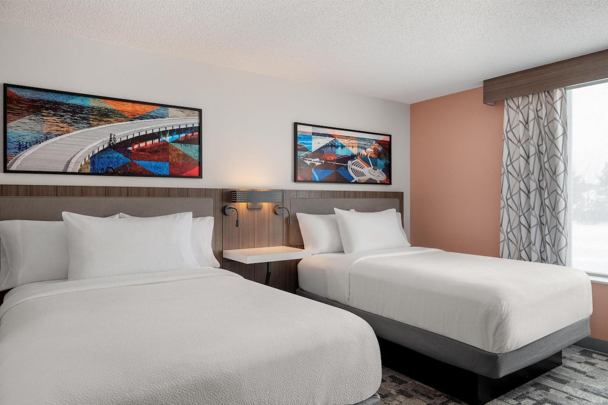 A hotel room with two beds, a headboard with built-in lighting, a large window with a view, and two framed artworks on the wall.