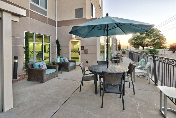 Une terrasse extérieure avec une table ronde et des chaises, un grand parasol et un coin salon avec des meubles en osier.