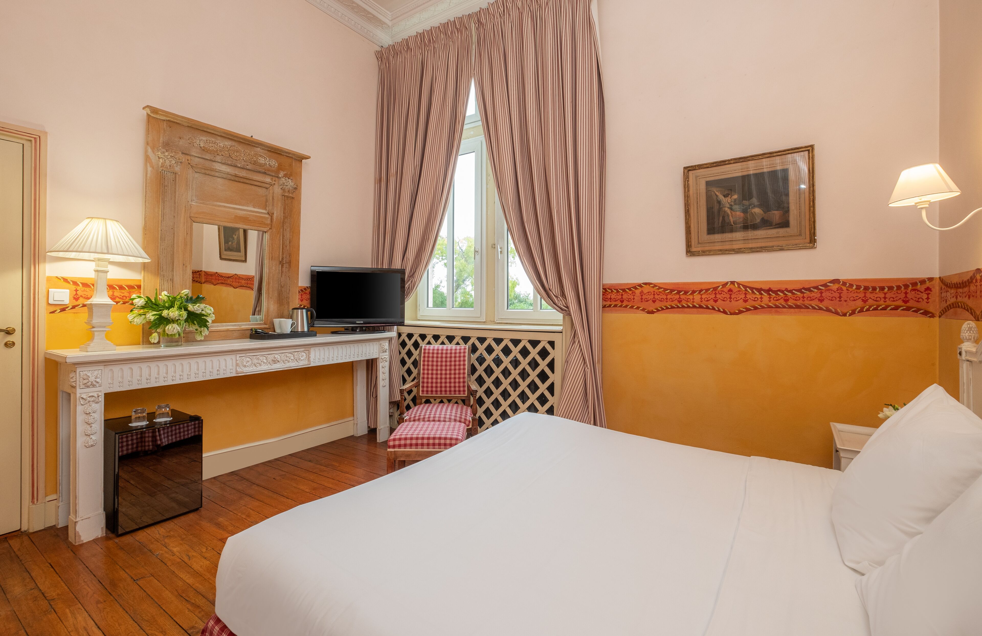 A hotel room with a bed, a desk with a computer, a chair, a window with curtains, a lamp, and a painting on the wall.