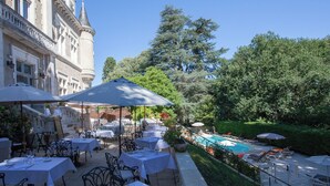 Een terras met tafels en parasols, een zwembad en weelderige begroeiing.