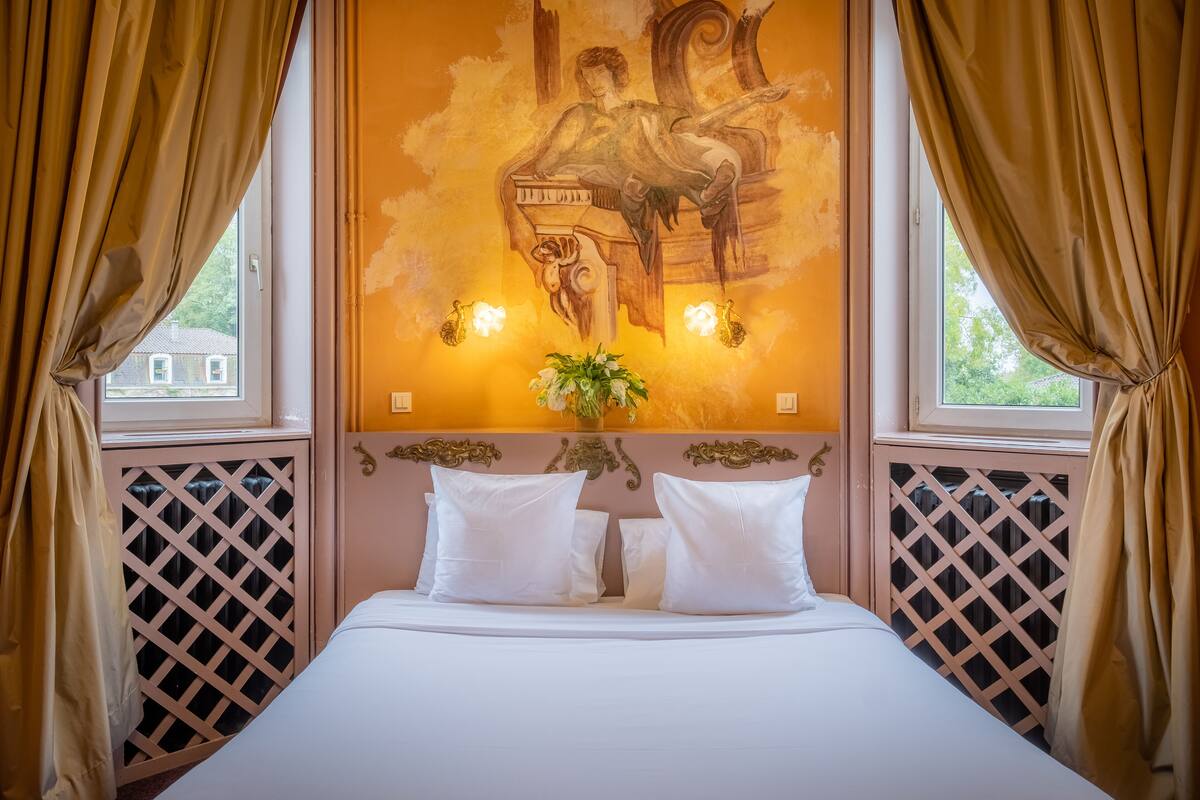 A bedroom with a bed, two white pillows, a vase with flowers, and a wall mural.