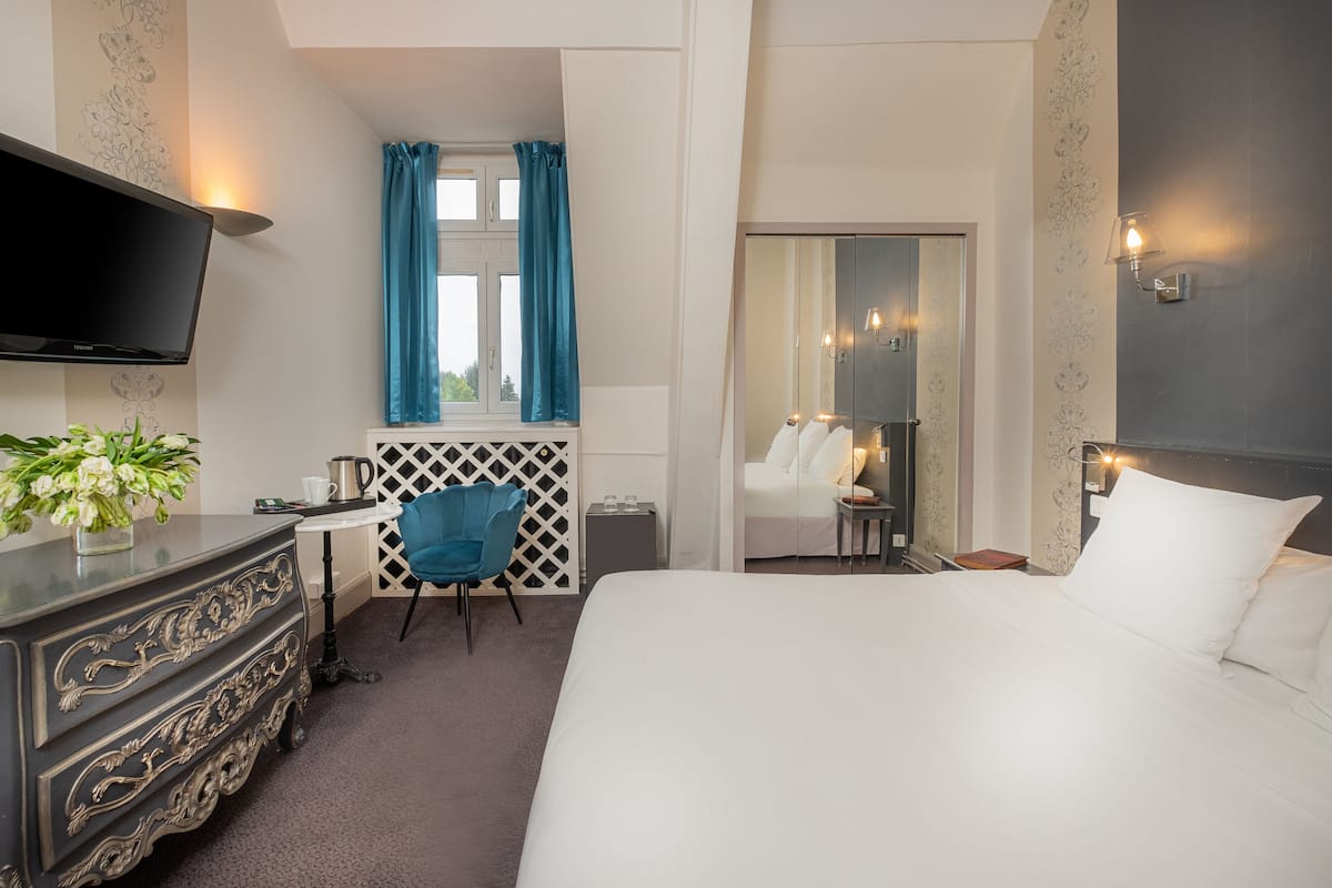 A hotel room with a large bed, a TV mounted on the wall, a desk with a chair, a small table with a lamp, and a mirror.