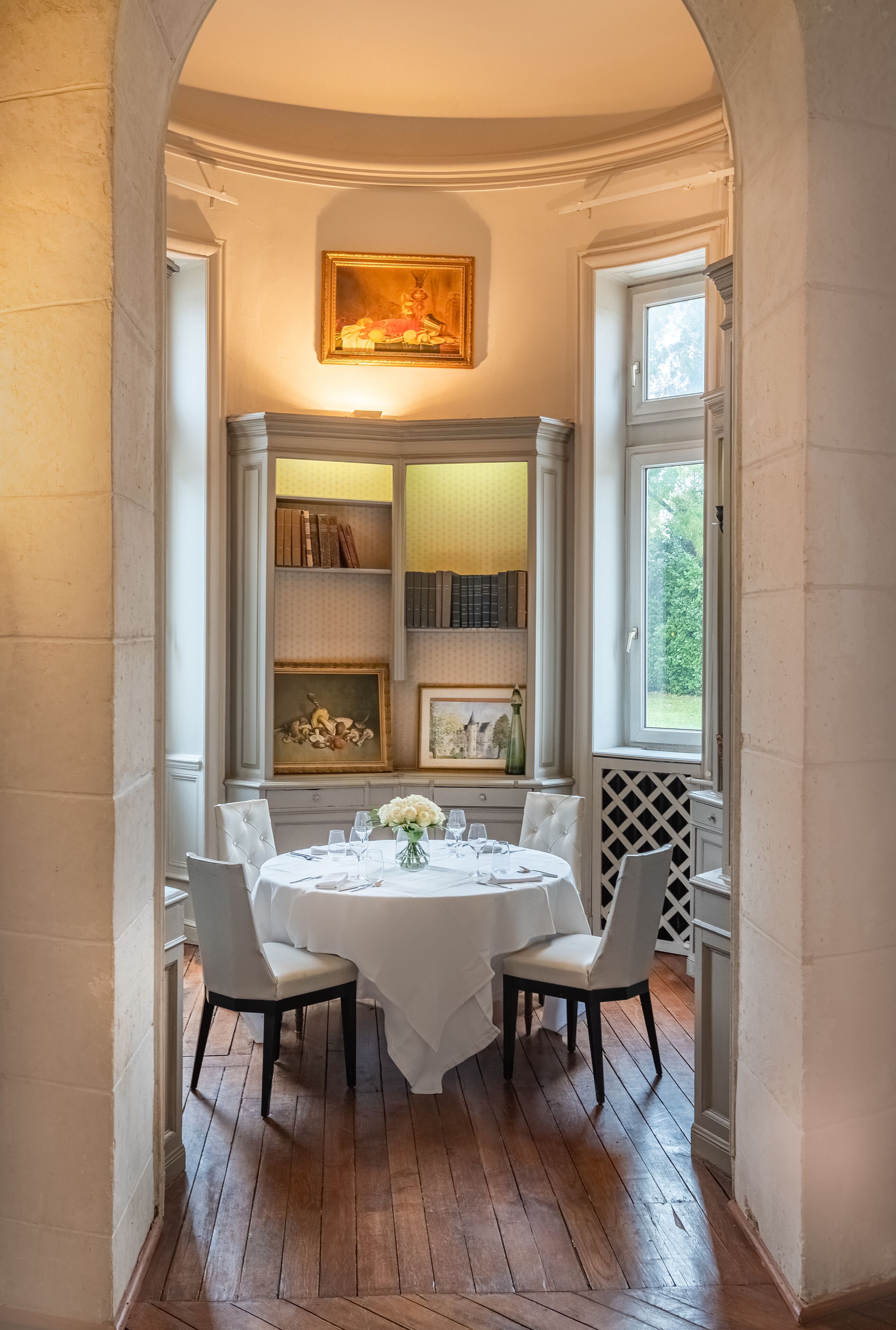 A dining area with a round table set for four, flanked by two chairs. A bookshelf with books and decorative items is mounted on the wall above the table.