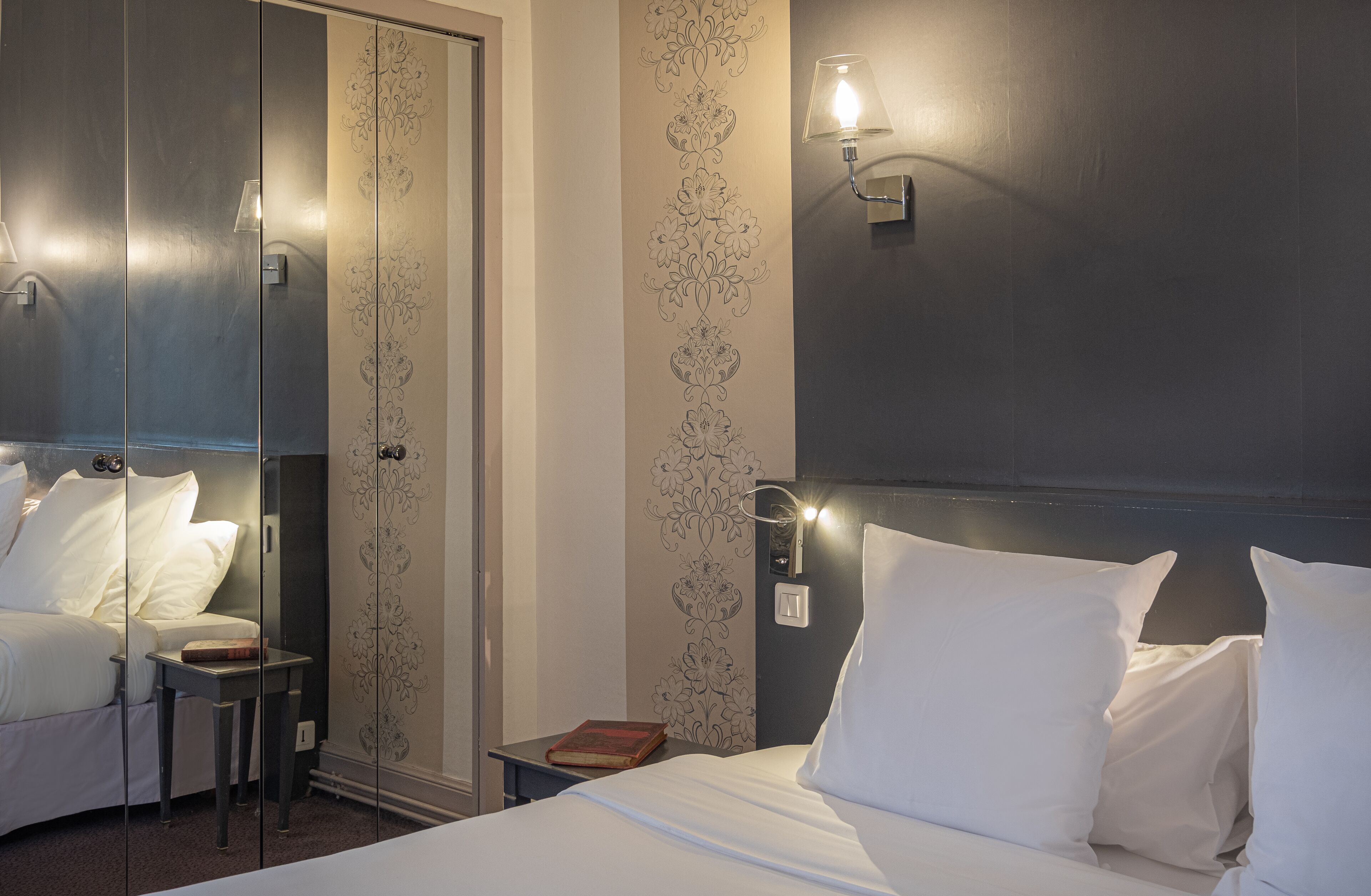 Une chambre d’hôtel comprenant un lit, des tables de chevet, une commode, un miroir, une armoire et une lampe fixée au mur.