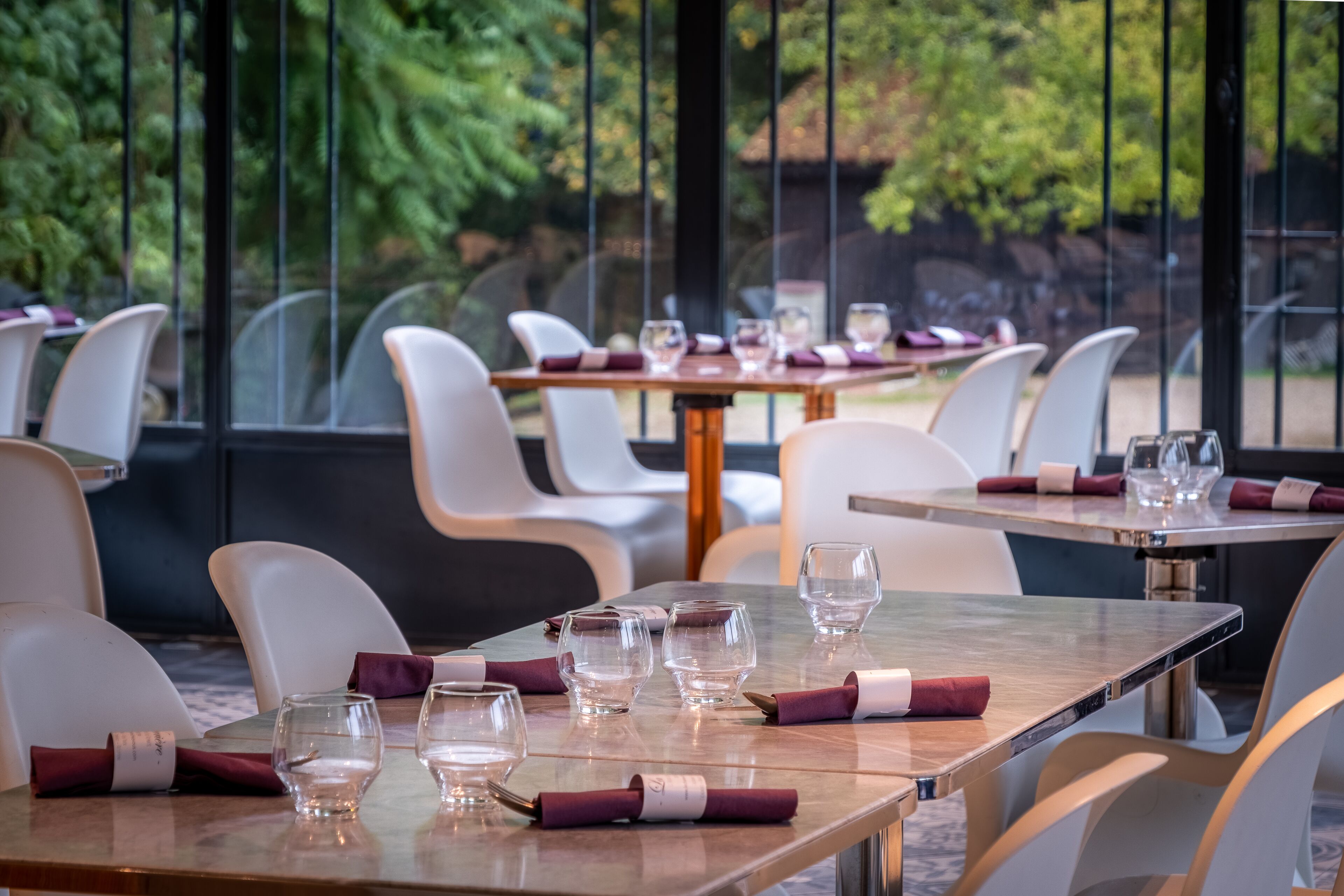 A restaurant with tables set for dining, chairs, and a view of trees through large windows.