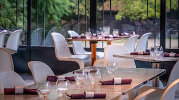 A restaurant with tables set for dining, chairs, and a view of trees through large windows.