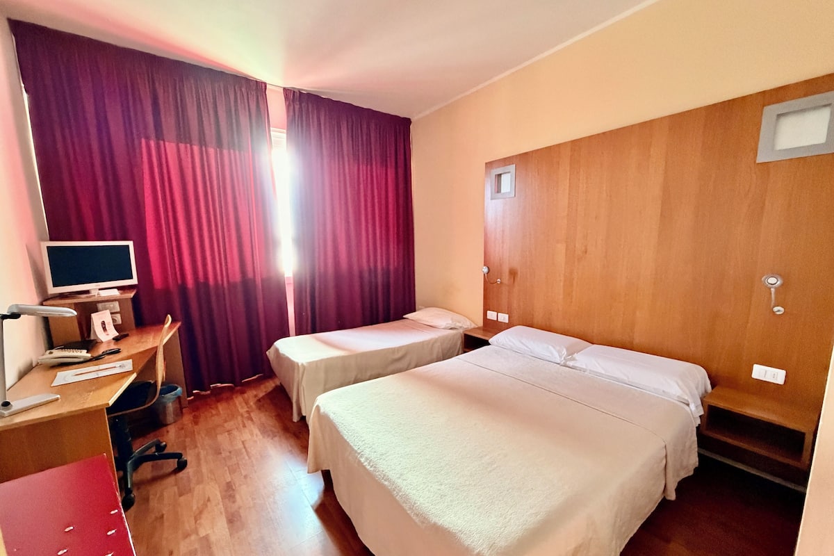 Una camera d'albergo con due letti, una scrivania con un computer e una finestra con tende.