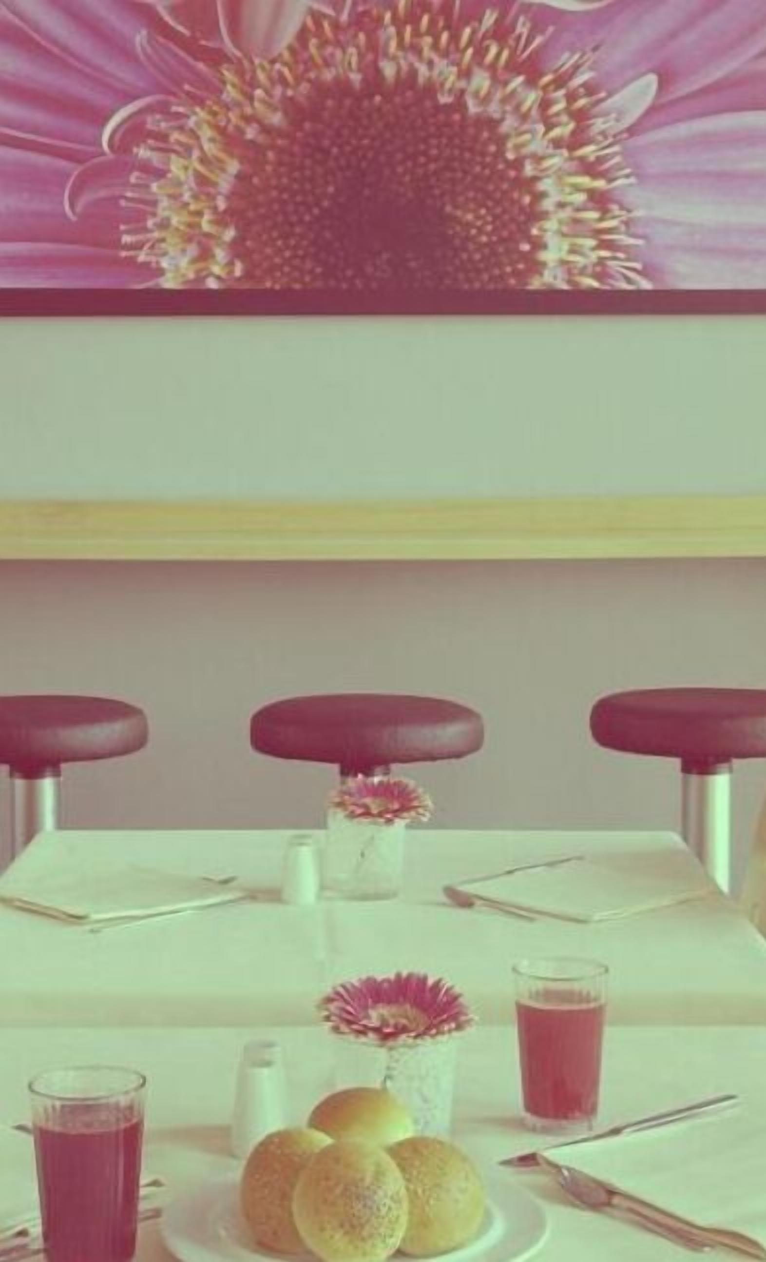 A dining table with three chairs, a vase with flowers, a plate with bread rolls, and two glasses of red drink.