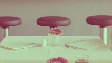 A dining table with three chairs, a vase with flowers, a plate with bread rolls, and two glasses of red drink.