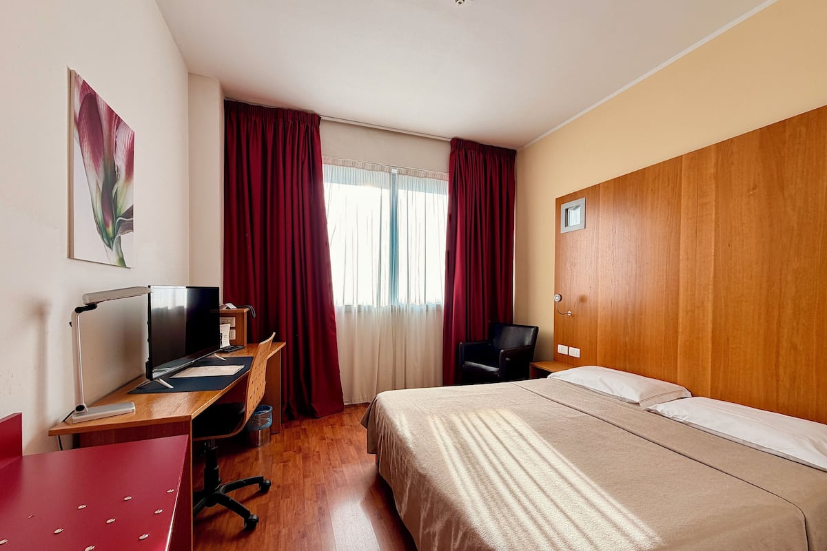 Una camera d'albergo con una scrivania in legno, un letto, una sedia e una finestra con tende.