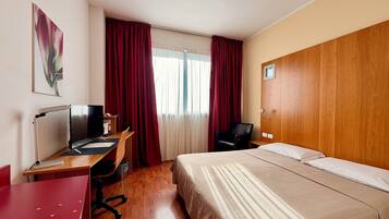 A hotel room with a wooden desk, a bed, a chair, and a window with curtains.