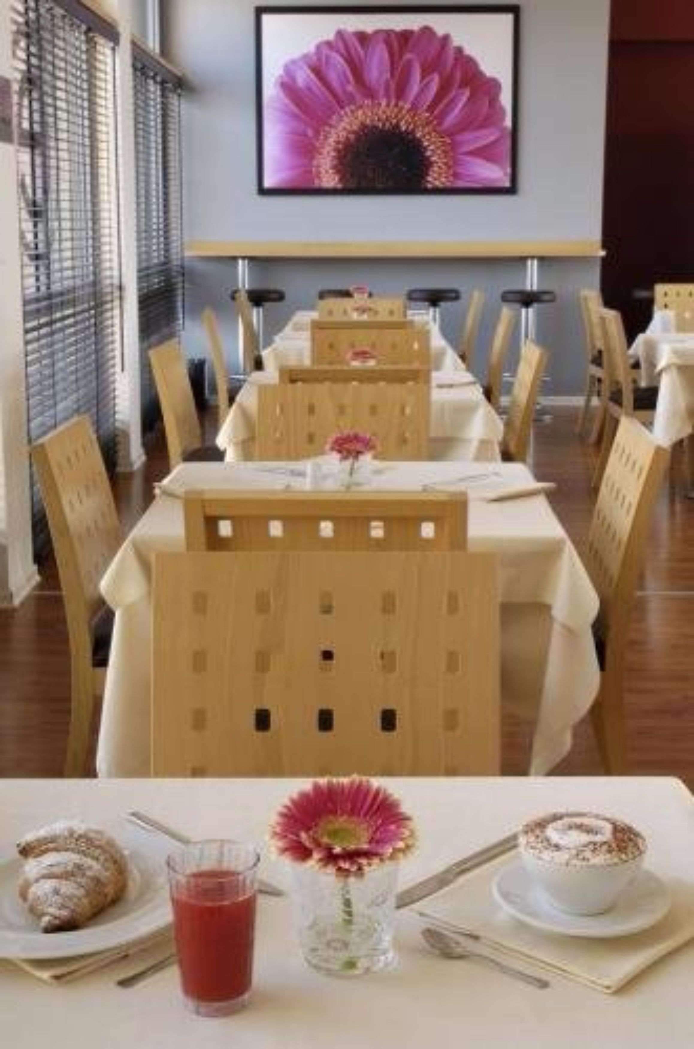 A dining area with wooden chairs and tables, a framed flower painting on the wall, and a table set with a croissant, a glass of red drink, and a coffee with whipped cream.