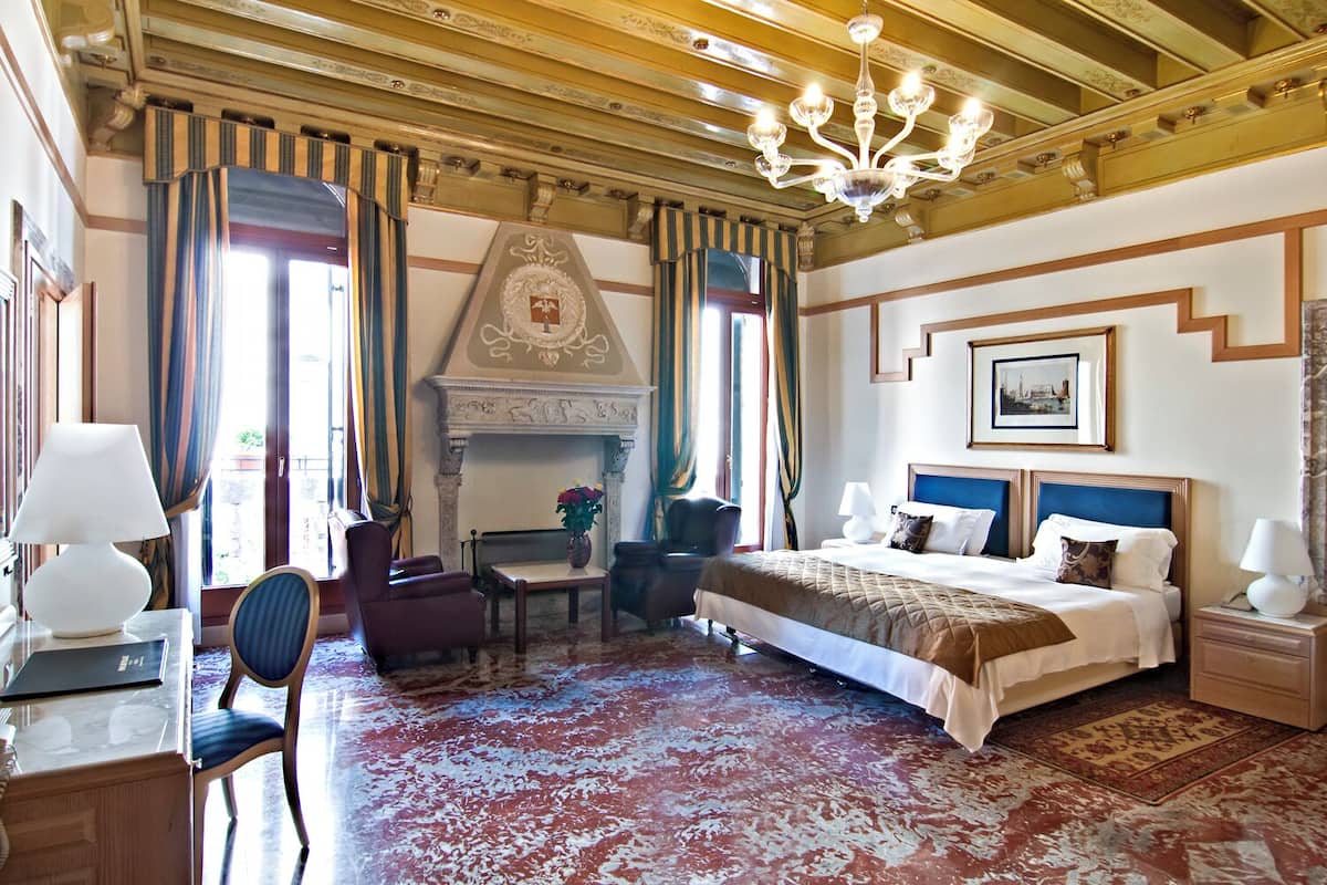 A hotel room with a large bed, a fireplace, a desk with a lamp, and a chair.
