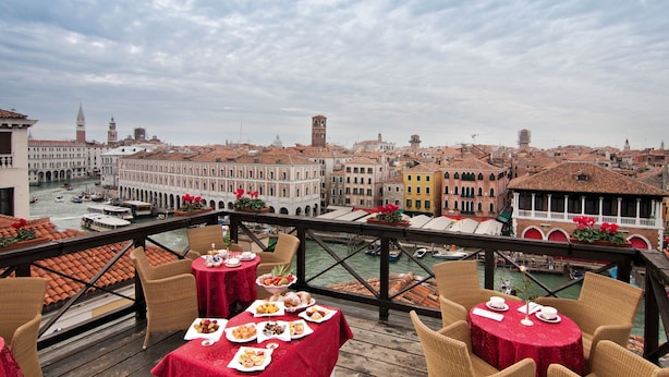 Terraza con mesas dispuestas para comer, con vistas a un canal y edificios históricos con barcos.