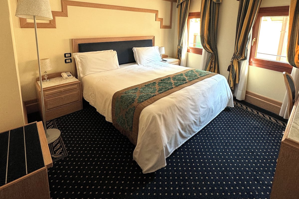 A hotel room with a bed, a nightstand, a lamp, a window with curtains, and a patterned carpet.