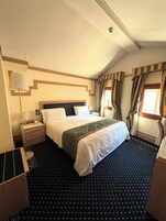 A hotel room with a bed, a nightstand, a lamp, a window with curtains, and a patterned carpet.
