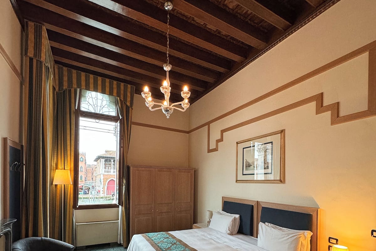 A hotel room with a large bed, a wooden ceiling, a view through a window, a nightstand, and a chair.
