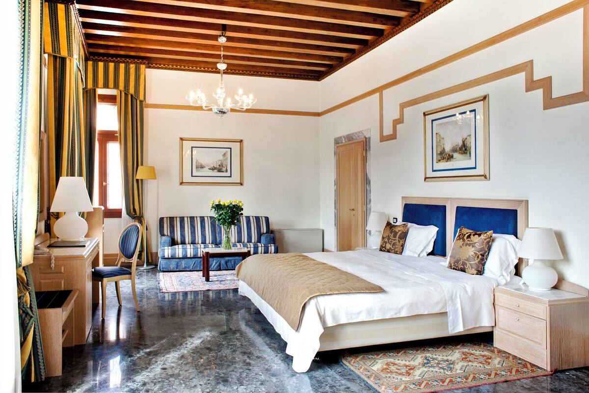 A hotel room with a large bed, a sofa, a desk, and a chair. There are two paintings on the walls, a chandelier, and a window with curtains.