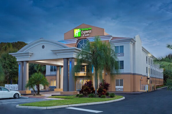 Um Holiday Inn Express & Suites com design moderno, apresentando entrada central com galeria coberta, palmeiras e estacionamento.