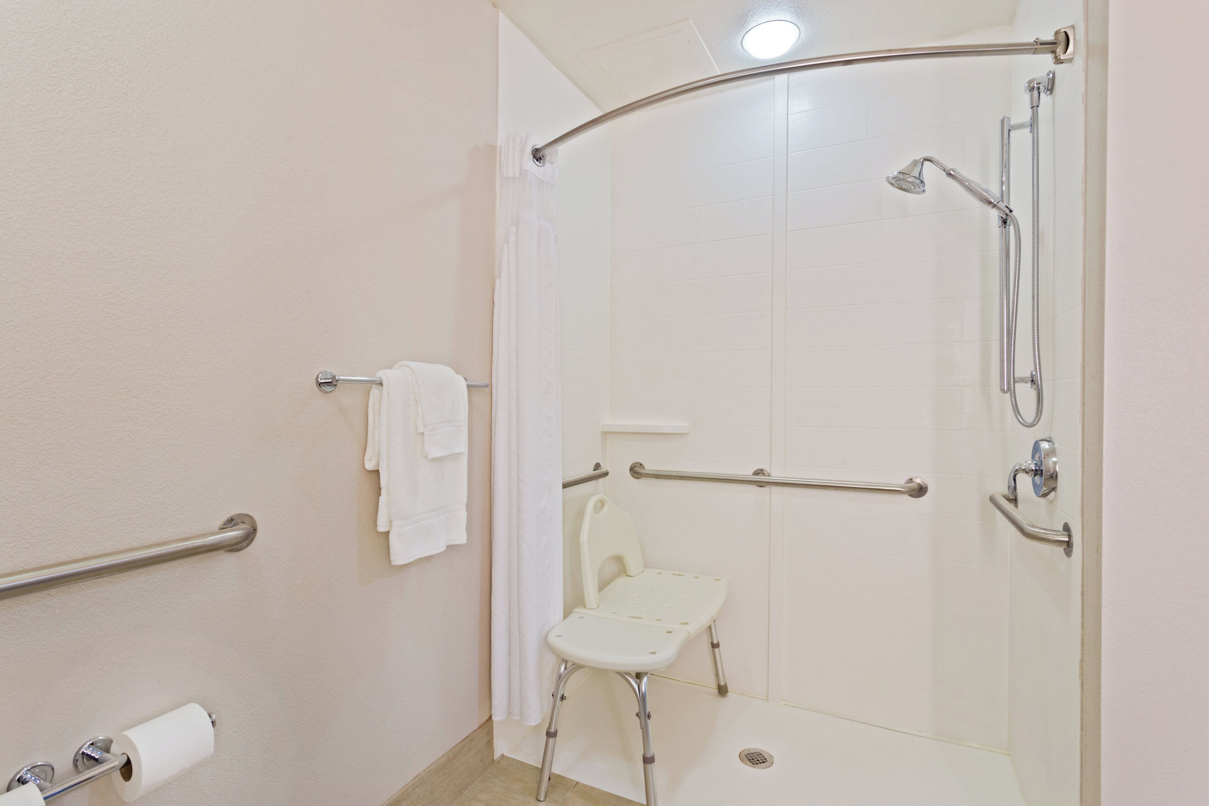 Et badeværelse med brusekabine, gelænder, toilet med sikkerhedssæde og et håndklædetørrestativ.