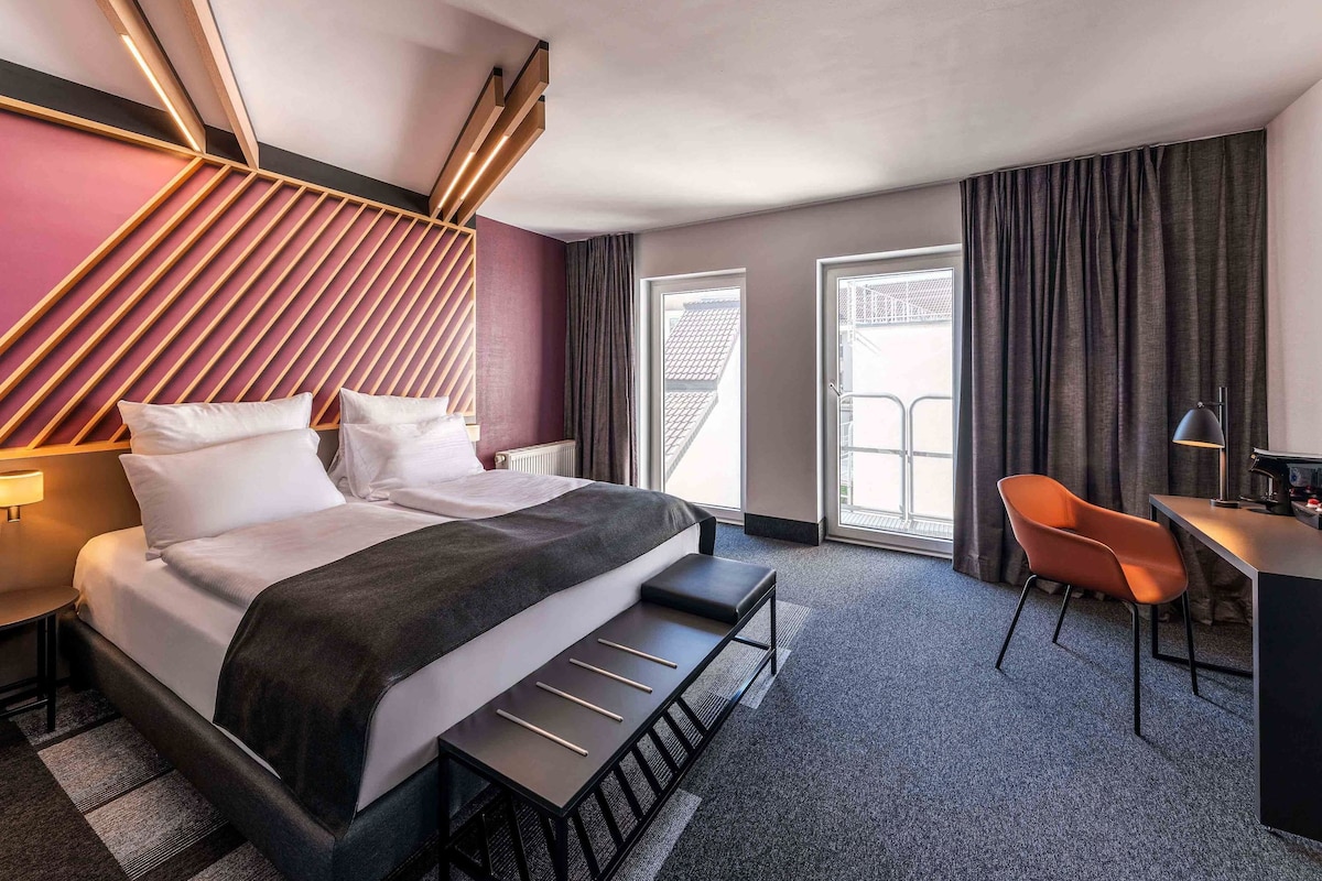 A modern hotel room with a large bed, a desk, and a chair. There are curtains on the windows and a view of the outside.
