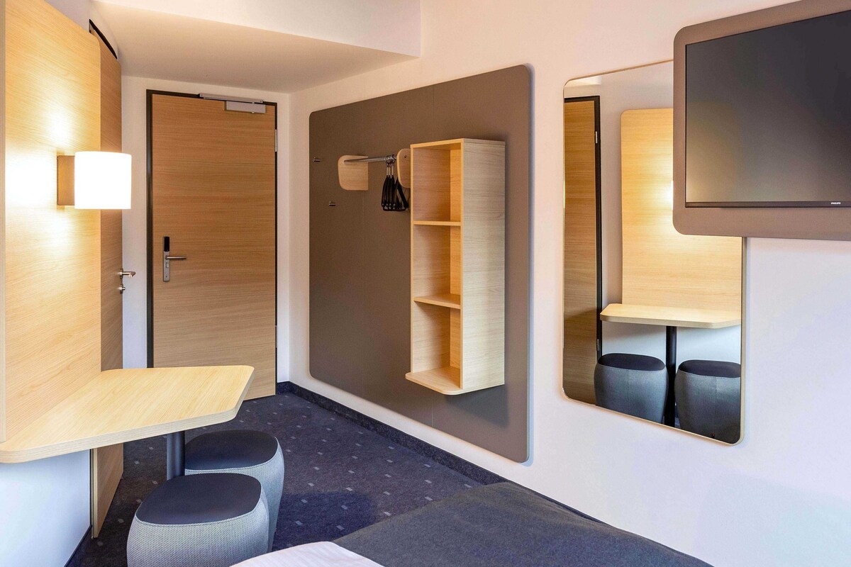 A modern hotel room with a wooden door, a small desk with a chair, a mirror, and a wall-mounted shelf.