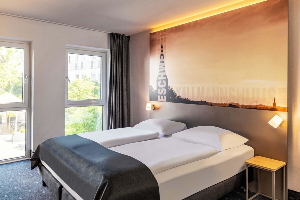 A hotel room with a bed, a small bedside table, a window with curtains, and a wall with a cityscape mural.