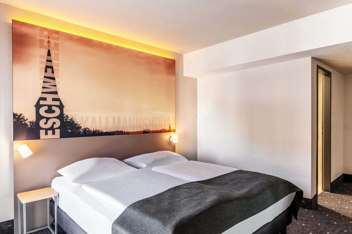A hotel room with a bed, a nightstand, a wall-mounted lamp, and a framed picture of a cityscape.