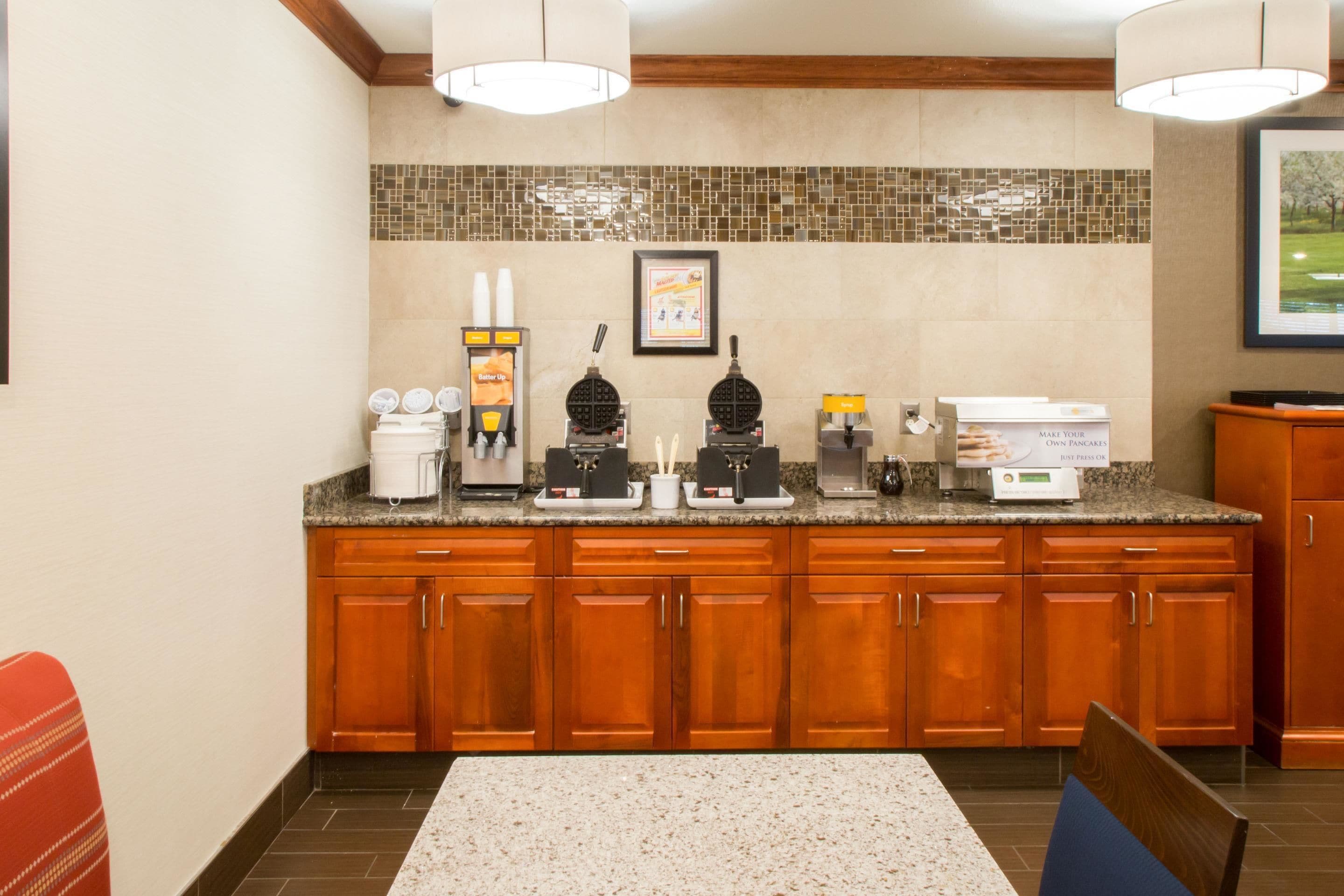 A hotel kitchen with wooden cabinets, a granite countertop, and various appliances including a waffle maker and coffee machines.