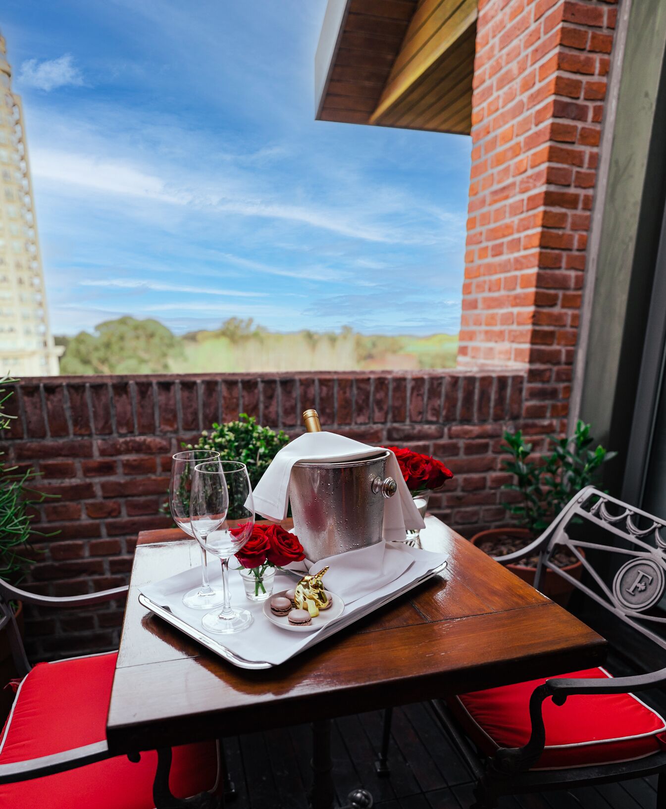 Une table dressée avec un seau à champagne, des verres, des roses rouges et des macarons, le tout posé sur une table en bois agrémentée d’un coussin rouge.