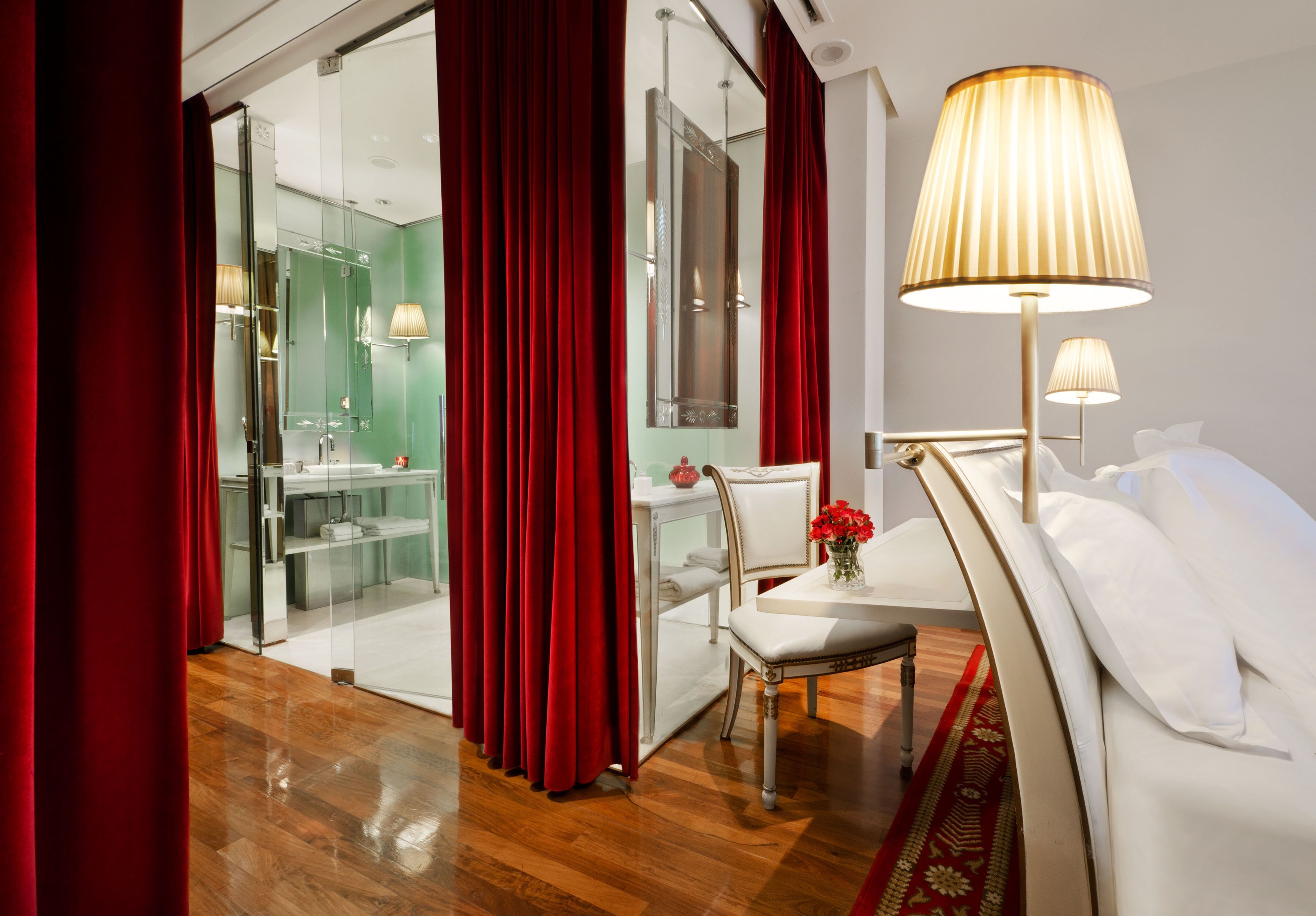 Habitación de hotel con piso de madera, cama blanca con almohadas, mesita de noche con lámpara, una silla y una pequeña mesa con un florero.
