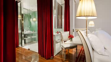 Habitación de hotel con piso de madera, cama blanca con almohadas, mesita de noche con lámpara, una silla y una pequeña mesa con un florero.