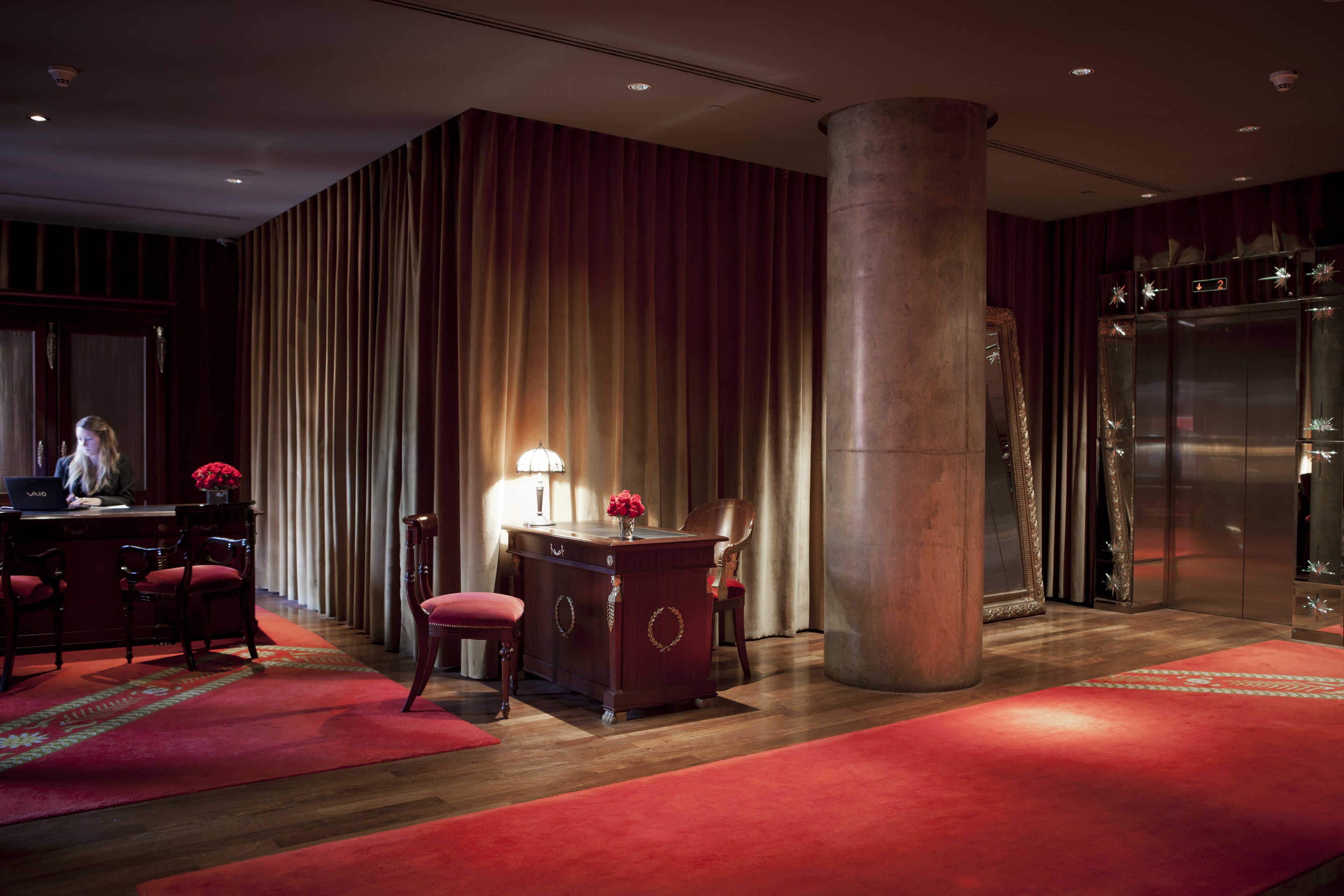 A hotel lobby with a reception desk, chairs, a large pillar, and a red carpet.