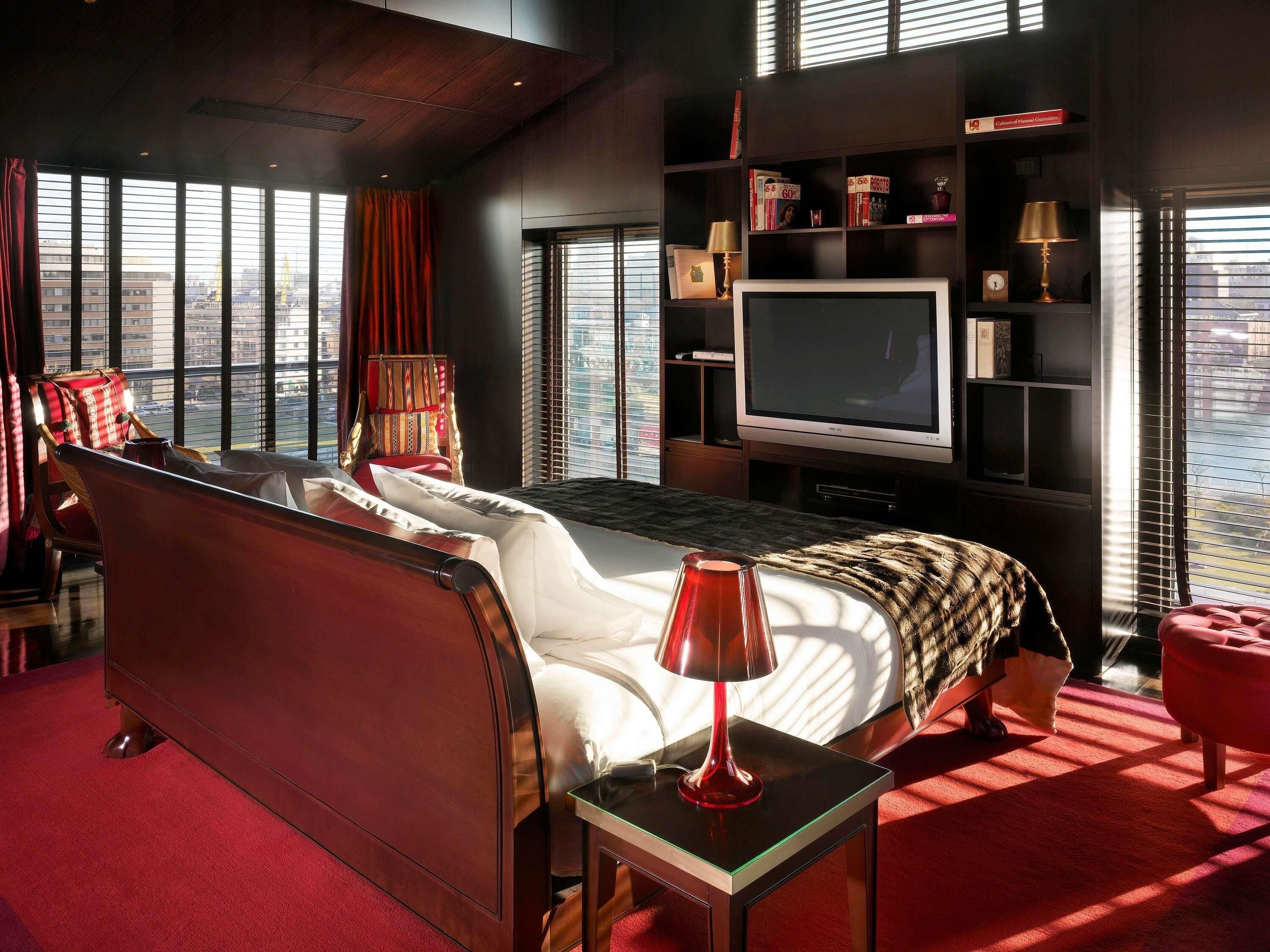 A modern hotel room with a large bed, a television, a bookshelf, and a view of the city through large windows.