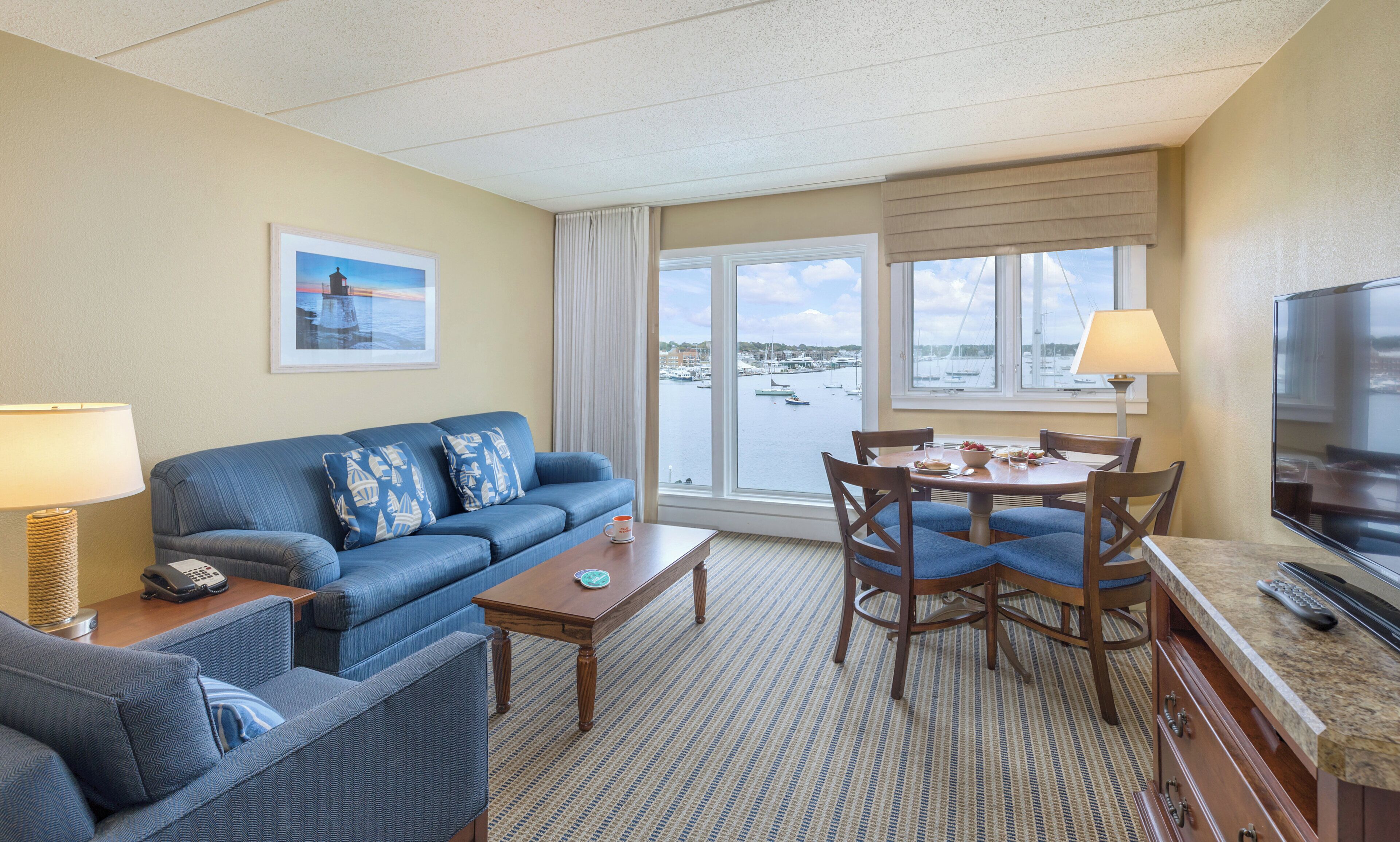 Une chambre d’hôtel dotée d’une grande fenêtre donnant sur les bateaux et l’eau, d’un canapé avec des coussins assortis, d’une table à manger avec des chaises, d’une télévision et d’une lampe.