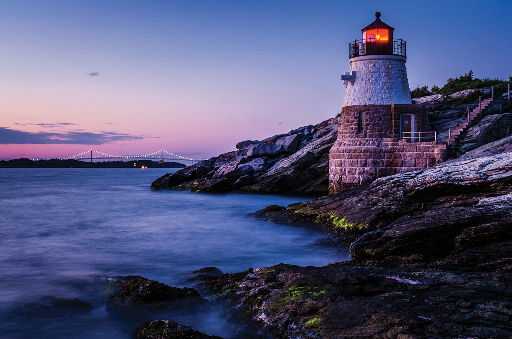 A lighthouse on rocky terrain with a bridge in the background.