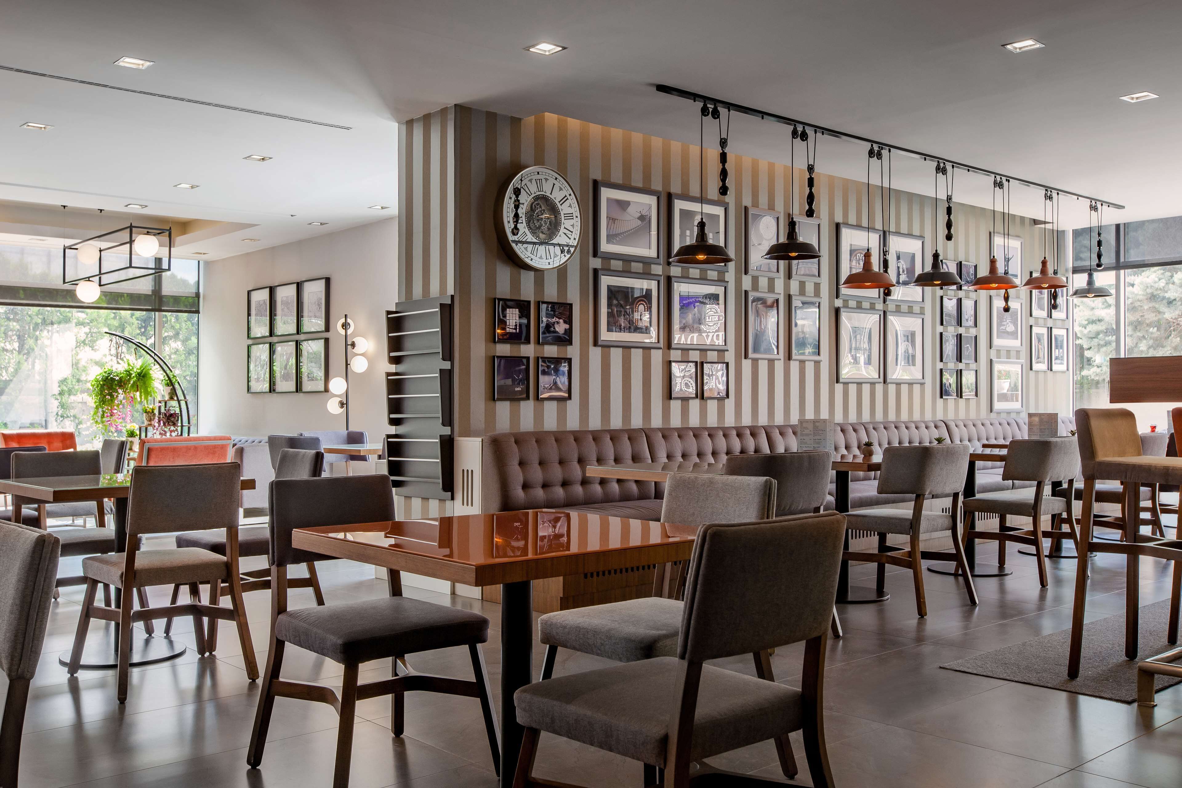 Um café moderno com paredes listradas, um relógio grande e quadros decorativos. Mesas e cadeiras dispostas para acomodar clientes.
