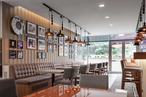A modern restaurant interior with a long bench seating area, multiple hanging pendant lights, framed pictures on the walls, and large windows offering a view of the outdoors.