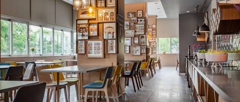 Un comedor moderno con mesas y sillas, cuadros en las paredes y amplios ventanales.