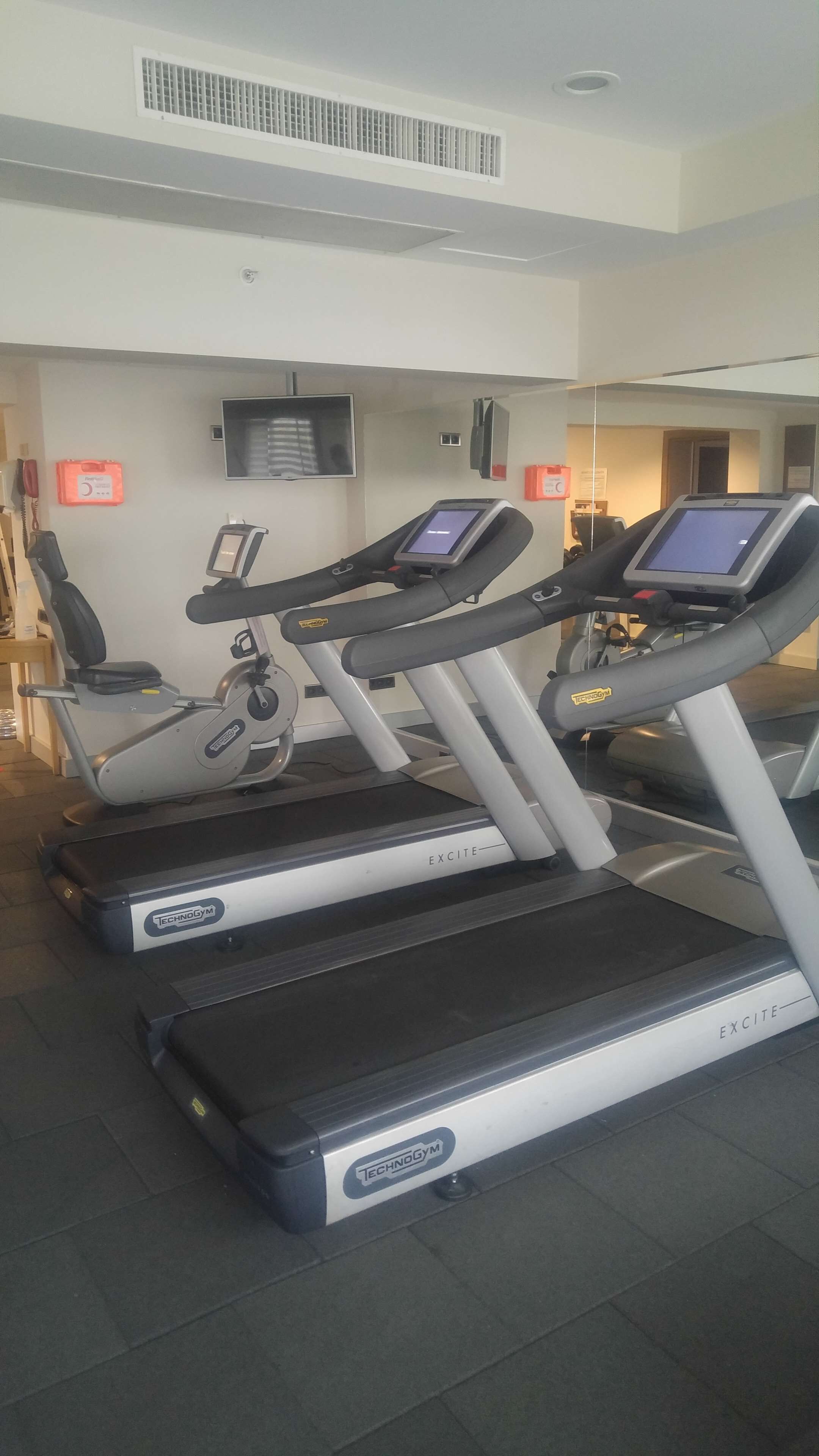 Un gimnasio con cintas de correr, un televisor en la pared y una señal de salida de emergencia a la vista.