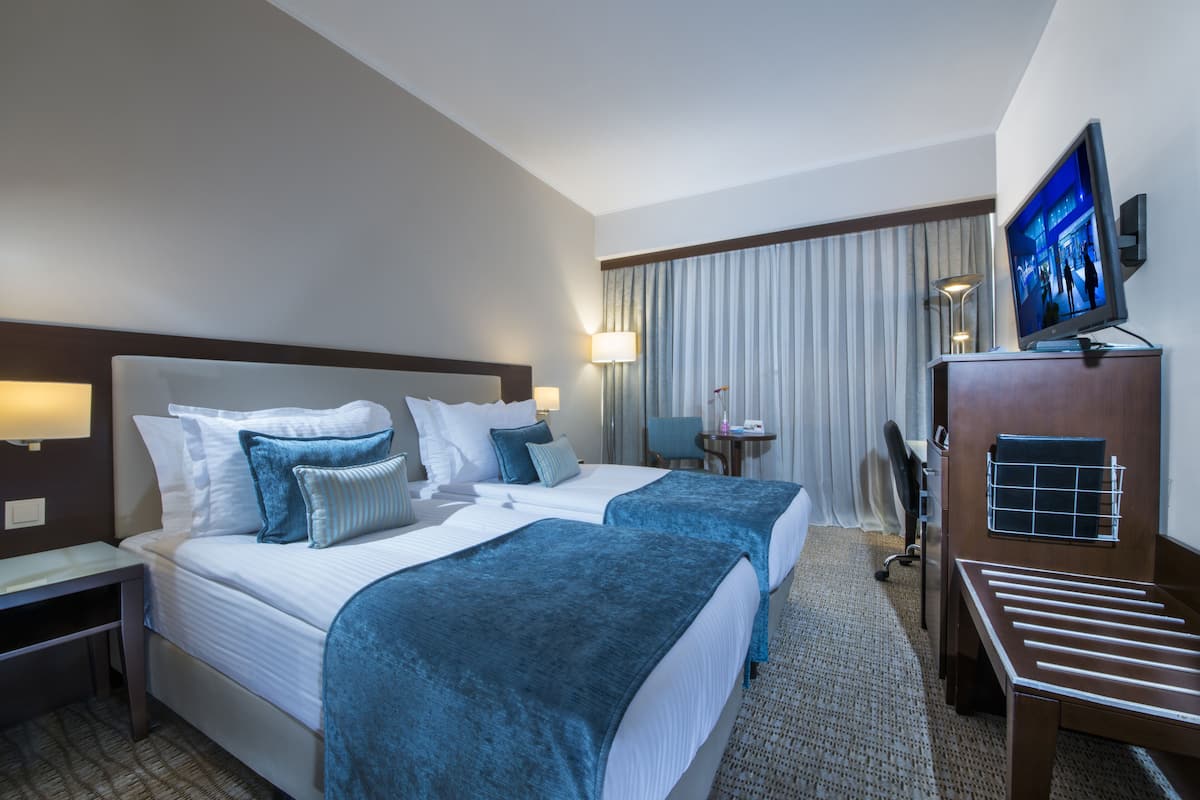A hotel room with two beds, a desk, a chair, a television, and a window with curtains.