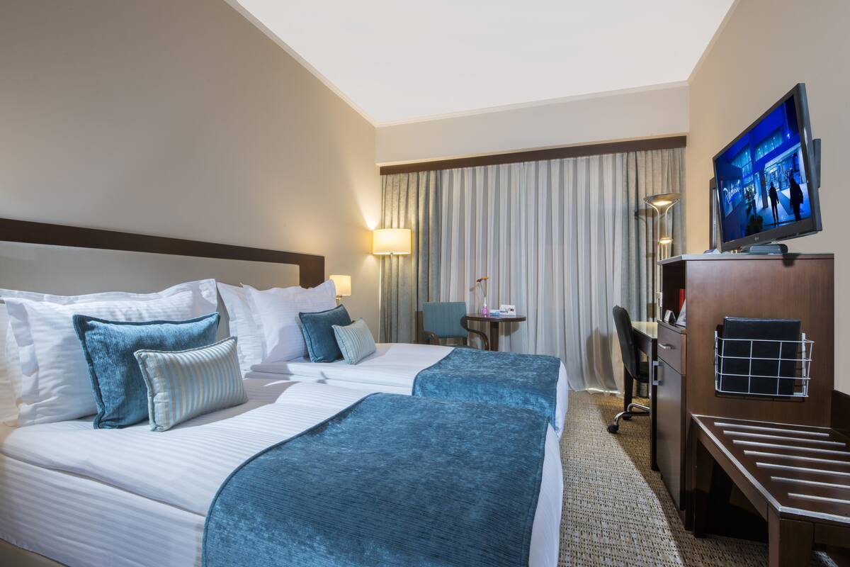 A hotel room with two beds, a desk, a chair, a TV, and a lamp.
