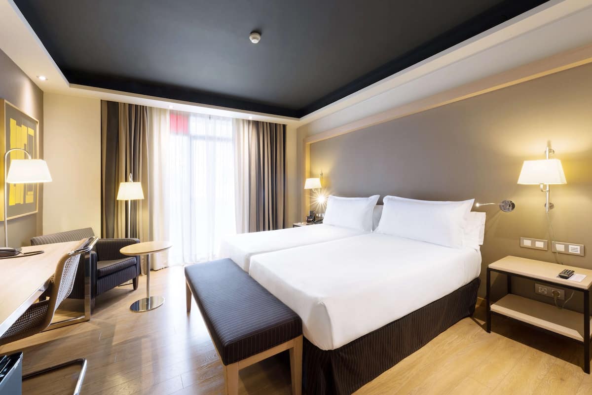 Habitación de hotel con una cama grande, dos mesitas de noche con lámparas, un escritorio con una silla, un banco y vistas al exterior a través de la ventana.
