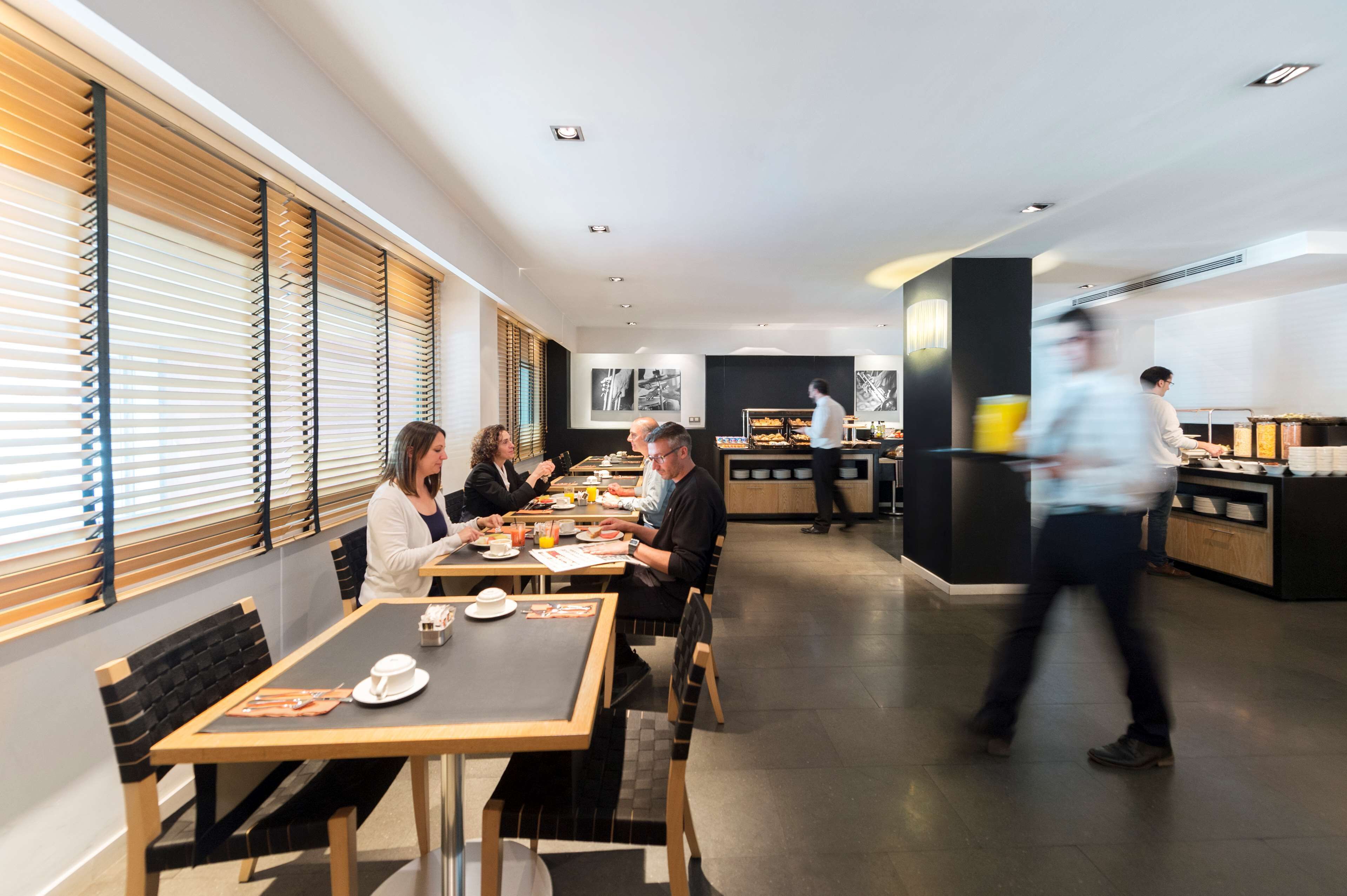 A modern dining area with wooden tables and black cushioned chairs, a buffet counter in the background, and a person walking by.